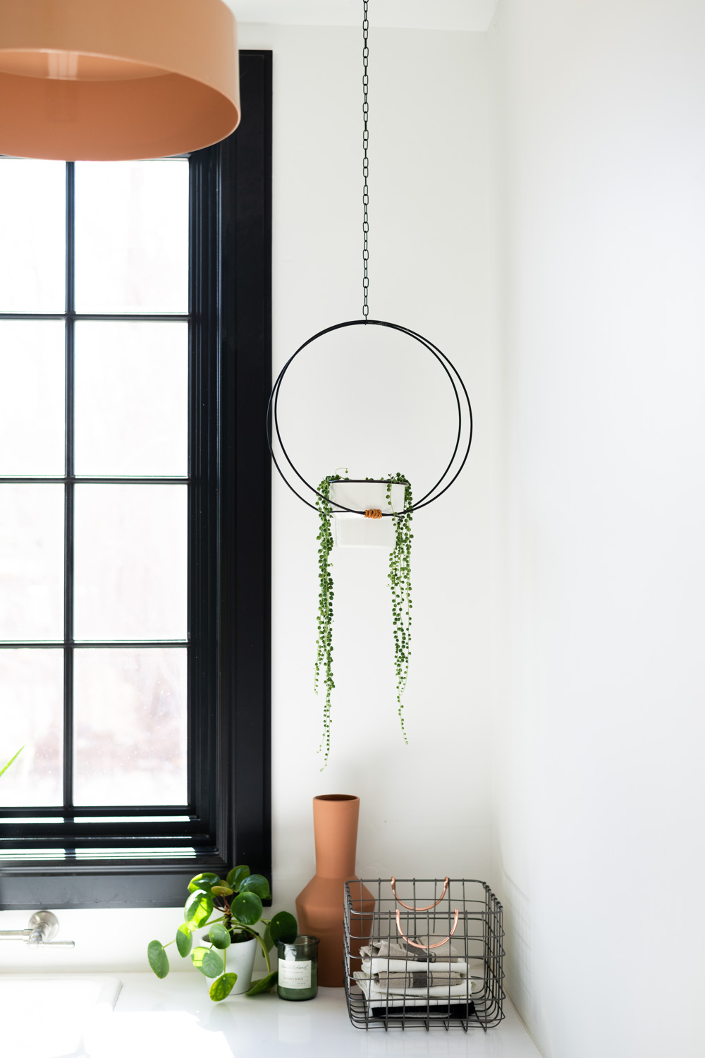 Black modern hanging planter with white pot and string of pearls succulent. Black window frame, wire basket with pilea and terra cotta vase